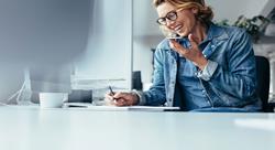 woman smiling in her office, jotting down notes while talking on her cellphone